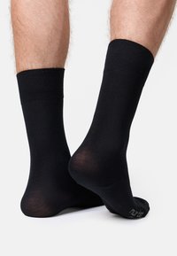 Schwarze Socken, die an den Füßen einer Person gezeigt werden, mit Nahaufnahmen der Stoffdetails und der gerippten Bündchen vor einem schlichten Hintergrund.
