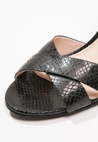 Sandale noire avec un motif brillant et texturé façon peau de serpent, avec des brides croisées et une semelle intérieure beige.
