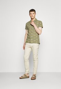 Kortærmet skjorte i gult og grønt geometrisk mønster, parret med lysebeige slim-fit bukser og grå sandaler med stropper.