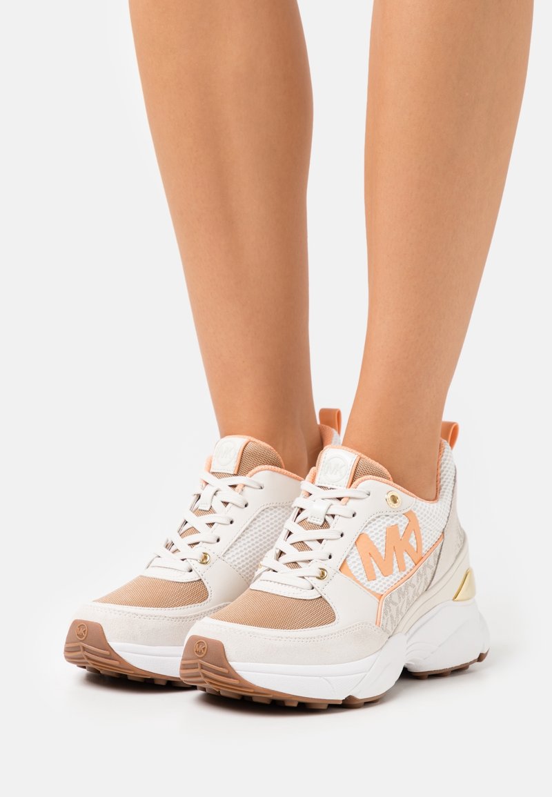 Baskets épaisses blanches et beiges avec des accents pêche et un grand logo MK portées aux pieds sur un fond uni.