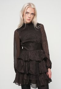 Γυναίκα με μακρύ πλατινέ ξανθό μαλλί, φορώντας μια διαφανή μαύρη μακρυμάνικη φόρεμα με στρώσεις και φρου φρου, μπροστά σε ένα απλό φωτεινό φόντο.