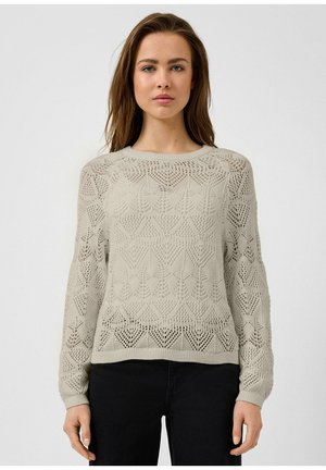 Giovane donna con indosso un maglione beige lavorato a maglia con un motivo geometrico traforato e pantaloni neri, in piedi davanti a uno sfondo chiaro e uniforme.