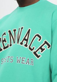 Felpa verde menta con scritte nere e beige, caratterizzata da un collo rotondo e una vestibilità comoda. Materiale in cotone resistente, tessuto morbido.