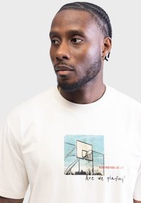 T-shirt bianca in cotone con grafica stampata di un campo da basket e testo "Are we playing?" e "REDEFINED REBEL EST. 2013" in colori a contrasto.