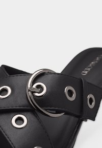 Μαύρο σανδάλι με φαρδιά λουράκι που διαθέτει ασημένιες μεταλλικές τρύπες και στρογγυλή ασημένια αγκράφα σε απλό φόντο.