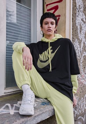 Juoda oversized marškinėliai su geltonu Nike logotipu, derinami su šviesiai žaliu džemperiu ir atitinkančiomis kelnių kelnėmis. Balti sportbačiai ir didelės auskarai.
