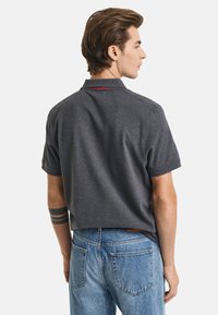 Graues Poloshirt mit kurzen Ärmeln, mit einem Kragen und einem dezenten roten Akzent auf dem Rücken. Kombiniert mit hellblauen Jeans.