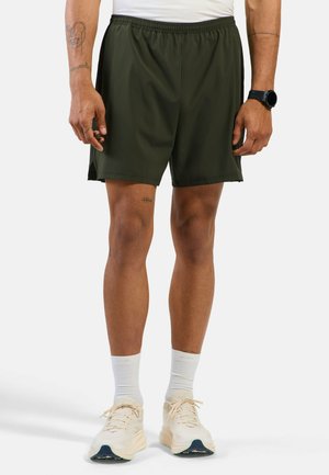 Mann trägt dunkelgrüne Sportshorts, weiße Socken und cremefarbene Laufschuhe, steht mit locker an den Seiten hängenden Armen.