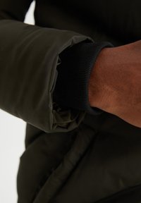 Mørk olivengrøn pufferjakkeærme med en sort ribbede manchet, der viser en glat tekstur og en let glans på stoffet.