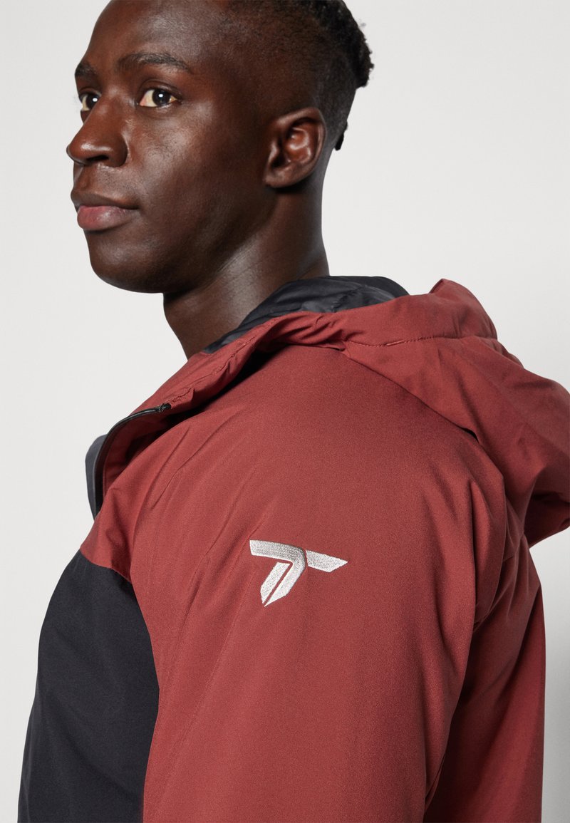 Uomo che indossa una giacca bicolore nera e rosso scuro con cappuccio e un logo geometrico bianco sulla parte superiore del braccio, che guarda di lato contro uno sfondo semplice.