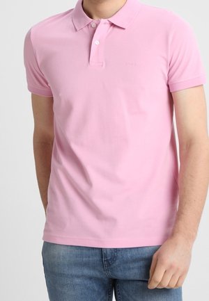 Homme portant un polo rose clair à manches courtes avec deux boutons et un jean bleu sur un fond uni.