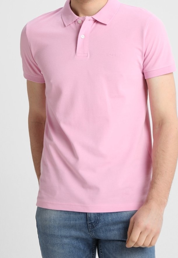 Homme portant un polo rose clair à manches courtes avec deux boutons et un jean bleu sur un fond uni.