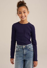 Hosszú ujjú lila bordázott póló, lekerekített nyakkivágással, világos kék farmernadrággal párosítva, amelynek rojtos szegélye és elülső zsebei vannak.