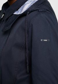 Zbliżenie na ramię granatowej kurtki z prążkowanym wykończeniem kołnierza, sznurkiem do ściągania oraz małą metalową tabliczką z logo na rękawie.