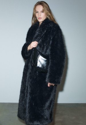 Sort faux fur frakke med høj krave og lange ærmer, båret over en sort læder crossbody taske, sat mod en neutral baggrund.