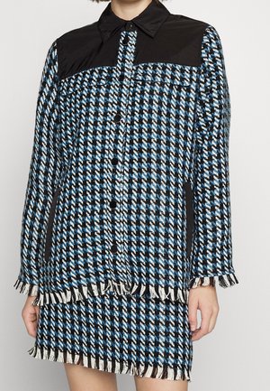 Modèle portant une veste bleu, noir et blanc à motif pied-de-poule, avec un empiècement noir et des franges, assortie à une jupe.