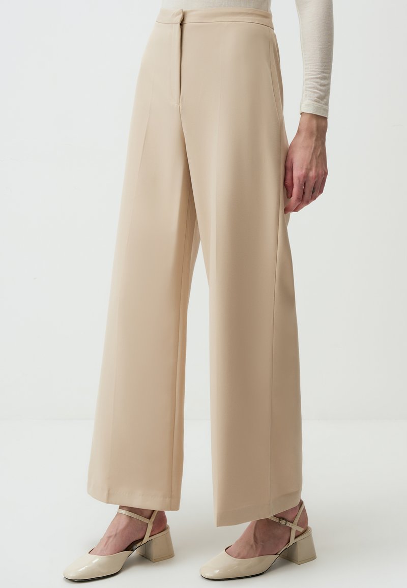 Beige wijde broekspijpen van smooth stof, met hoge taille, plooien en een nette afwerking. Gecombineerd met beige hakken.