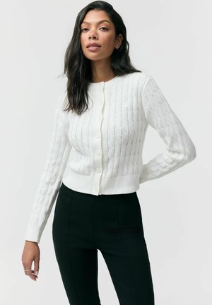 Kobieta o długich ciemnych włosach nosi biały, zapinany na guziki sweter w sploty oraz czarne spodnie z wysokim stanem, stoi z jedną ręką na biodrze.