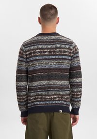 Mand set fra ryggen iført en multifarvet stribet uldsweater og olivengrønne bukser mod en ensfarvet hvid baggrund.