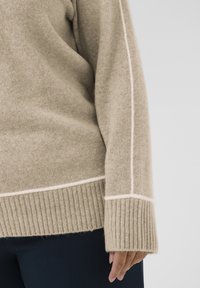 Pull en tricot beige avec poignets et ourlet côtelés, présentant une ligne blanche contrastante le long des coutures, en laine douce texturée.