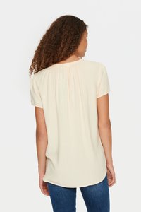 Blusa a maniche corte color crema chiaro con un motivo texturizzato e scollatura arricciata, indossata con jeans blu. Tessuto morbido e fluente.