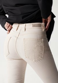 Jeans color beige claro con un diseño ajustado, bolsillos con parche texturizados y una etiqueta en la cinturilla. La tela parece suave con un ligero estiramiento.