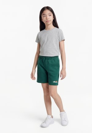 Joven con cabello largo y oscuro, vistiendo una camiseta gris, pantalones cortos verdes de Ellesse y zapatillas blancas, de pie contra un fondo blanco.