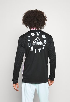 Osoba o kręconych włosach nosi czarną koszulkę Adidas z długim rękawem z "Love Unites" wokół logo na plecach, w zestawieniu z jasnymi spodniami.