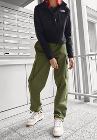 Chaqueta corta de forro polar negra con cremallera, pantalones cargo verdes con bolsillos laterales y zapatillas beige con detalles, sobre un fondo de buzones.