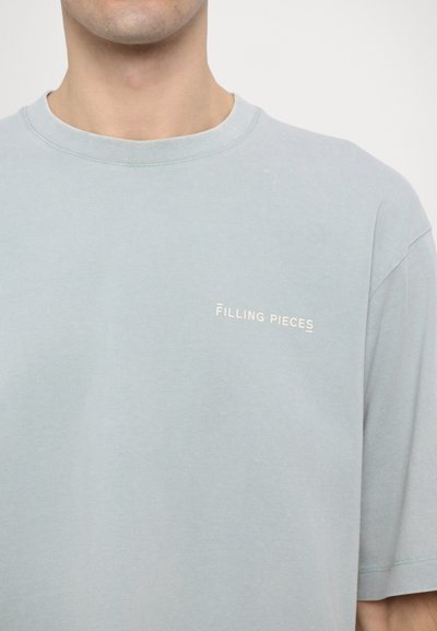 Světle modré bavlněné tričko s kulatým výstřihem, krátkými rukávy a malým bílým logem "FILLING PIECES" na levé části hrudi. Měkká textura.