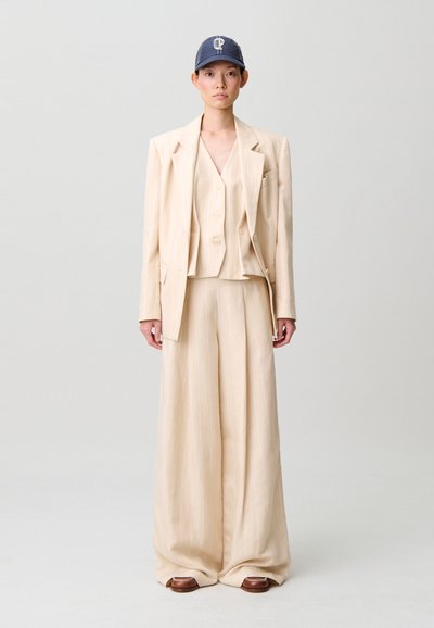 Completo beige a tre pezzi con righe sottili, composto da blazer su misura, gilet e pantaloni a gamba larga. Il modello indossa un berretto blu navy e scarpe marroni.