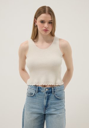 Joven mujer con cabello rubio y liso, lleva un top de punto sin mangas de color blanco roto y jeans azules de tiro alto, de pie contra un fondo neutro.