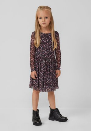 Junges Mädchen mit langen blonden Haaren, das ein schwarzes, blumengemustertes Langarmkleid und schwarze schnürbare Ankle Boots trägt, steht vor einem grauen Hintergrund.