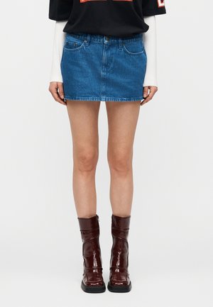 Persona que lleva una minifalda vaquera azul, camiseta de capas en blanco y negro, y botas de tobillo brillantes de color rojo oscuro, de pie frente a un fondo liso.