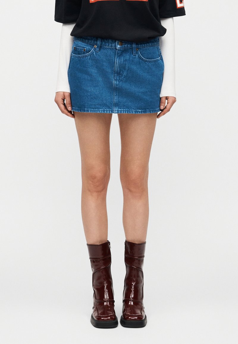 Person iført en blå denim miniskjørt, svart-hvitt lagdelt topp og skinnende mørkerøde ankelstøvler, stående mot en ensfarget bakgrunn.