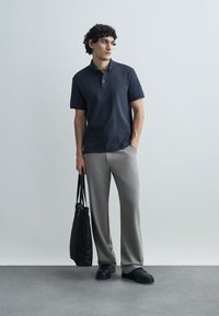 Μπλε ναυτικό πόλο με διακριτικές ριγέ υφές, συνδυασμένο με ανοιχτό γκρι παντελόνια. Το μοντέλο κρατά μαύρη τσάντα tote, φορώντας μαύρα αθλητικά παπούτσια.