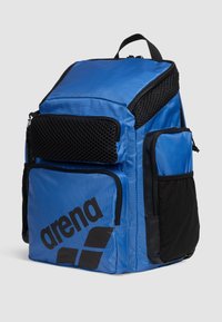 Mochila azul con múltiples compartimentos, hecha de tela duradera. Presenta un bolsillo de malla y acentos en negro contrastantes con un logo.
