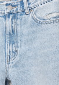 Jean en denim bleu clair en gros plan montrant la taille, le bouton en métal, la boucle de ceinture et un détail effiloché sur le bord de la poche avant.
