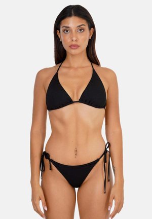 Femme aux longs cheveux foncés portant un bikini string noir avec des côtés noués, debout devant un fond clair uni.