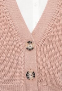Růžový pletený kardigan s výstřihem do V, s dvěma kulatými vícebarevnými knoflíky a strukturou ve vzoru pletení. Pod ním je bílá košile.