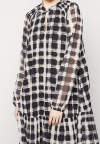 Robe à carreaux noir et crème avec un col rond, un lien de serrage, des manches longues et une jupe à volants en tissu léger et transparent.