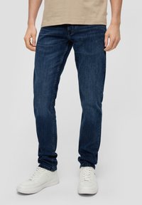 Tamno plavi traperice od denima, slim fit kroja, s pet džepova i petljama za pojas, uparene s bijelim tenisicama. Jednostavan dizajn i tekstura.