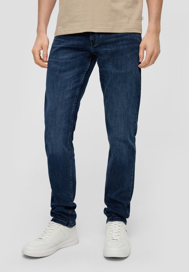 Tamno plavi traperice od denima, slim fit kroja, s pet džepova i petljama za pojas, uparene s bijelim tenisicama. Jednostavan dizajn i tekstura.