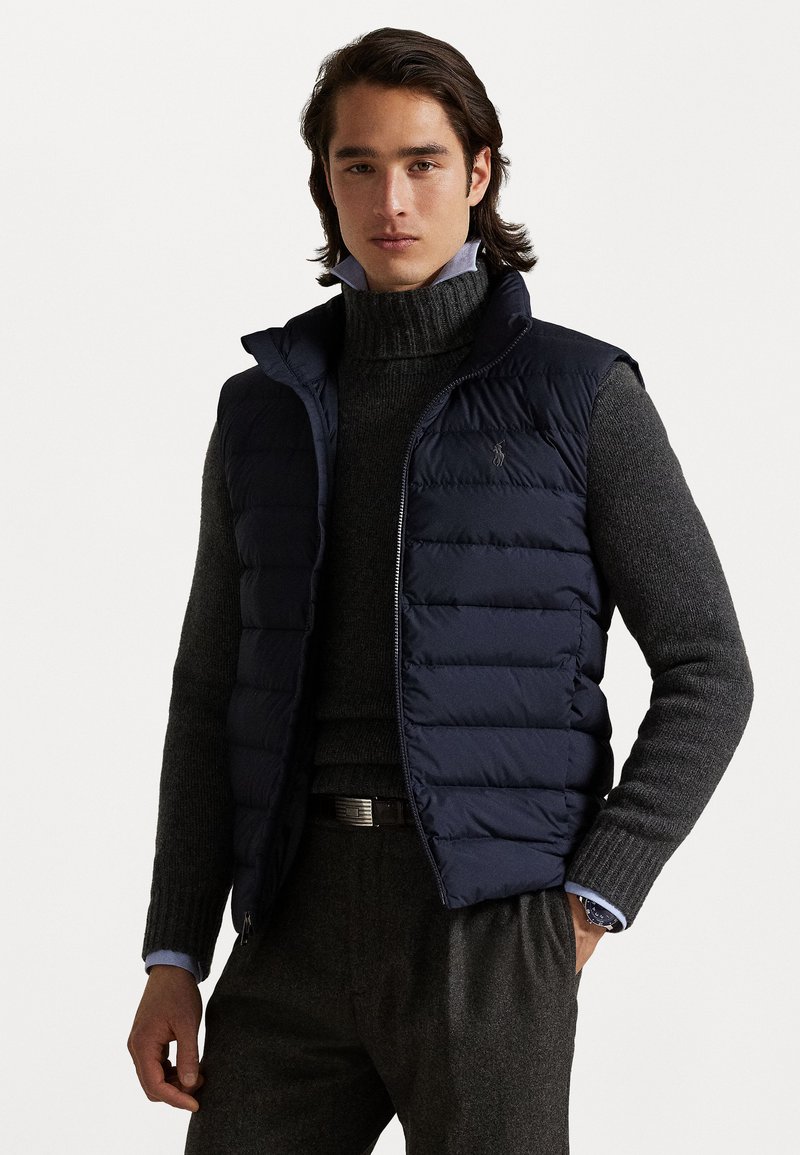 Navy blue padded vest with high collar, zip front, and logo accent. Worn over a dark gray turtleneck sweater, featuring ribbed texture.
