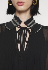 Femme portant un chemisier noir avec un col plissé haut avec des coutures blanches et noué avec des rubans noirs, assorti à de grandes boucles d'oreilles dorées.