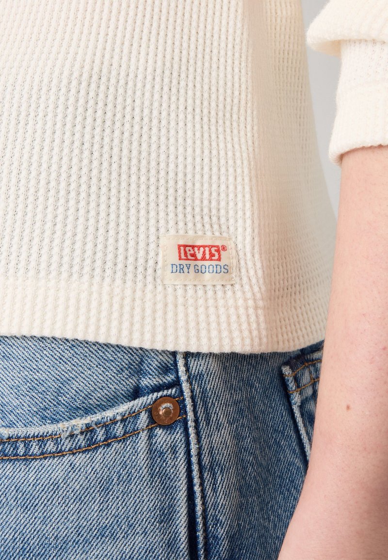 Suéter de canalé crema con una etiqueta de Levi's en letras rojas y azules, combinado con jeans de mezclilla azul claro, mostrando una vista en primer plano.