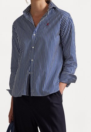 Blå og hvid lodret stribet skjorte med knapper og op rullede ærmer, båret med sorte bukser, hånd i lommen.