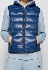 Person som bär en blank blå puffväst med dragkedja och fickor över en ljusblå hoodie och matchande sweatpants.