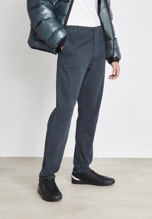 Pantaloni di cotone dritti di colore blu scuro con chiusura a bottone, abbinati a un giubbotto imbottito lucido e sneakers nere con un accento di colore.