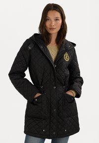 Svart vadderad jacka med huva, snörliv och två framfickor. Har ett dekorativt emblem på bröstet och strukturtyg.